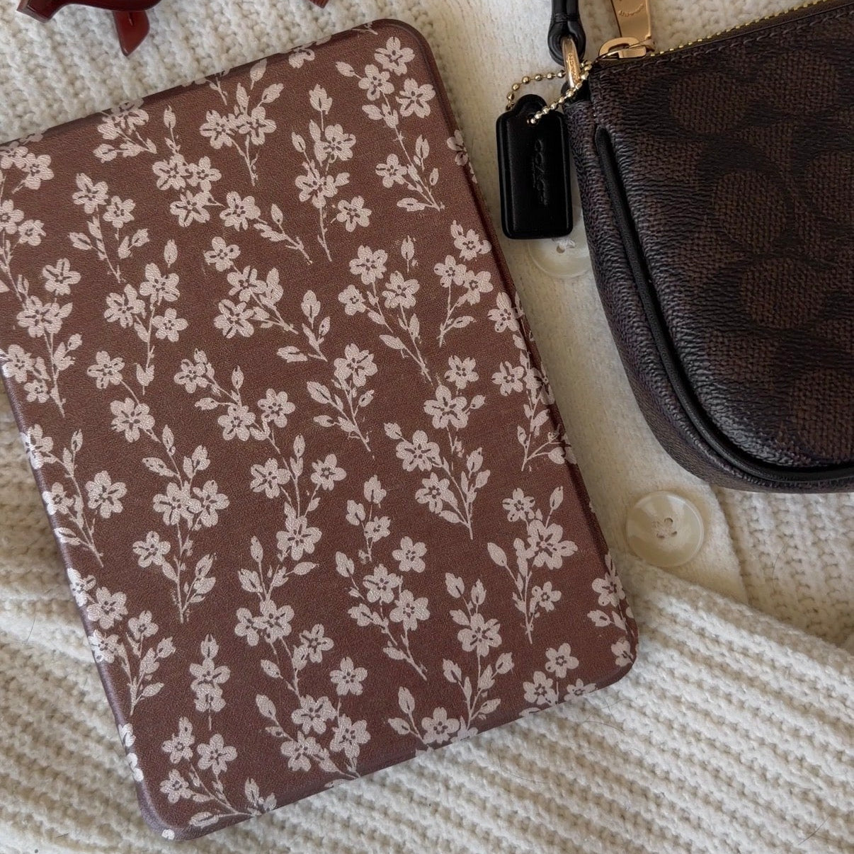 A styled flat-lay featuring a brown notebook with a white floral pattern, a brown eyeglasses case, a brown lip gloss tube, and a brown patterned wristlet bag, all arranged on a cream-colored knit blanket.